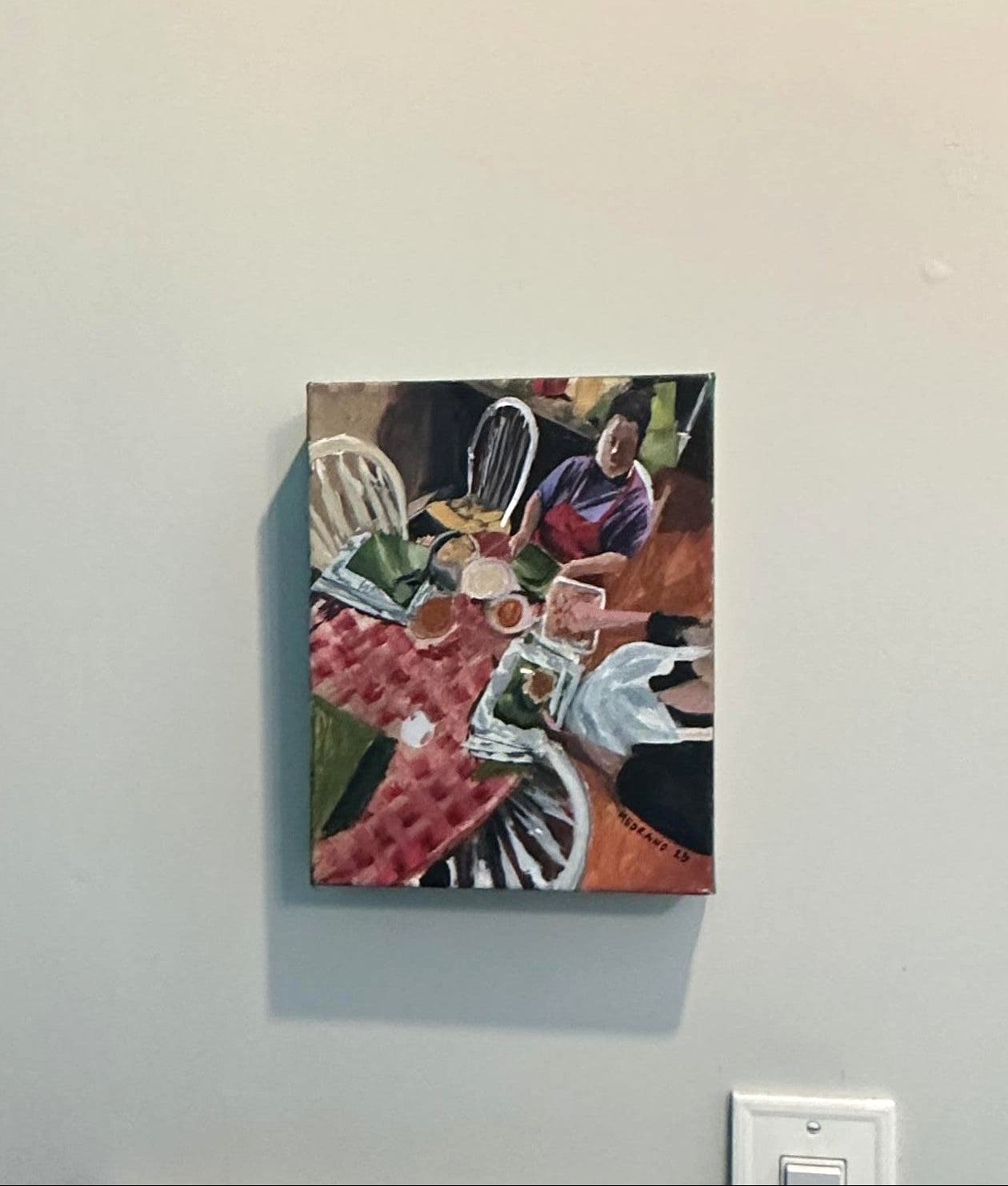 A small colorful painting depicting a table scene with people and objects, mounted on a light gray wall next to a teal door frame.